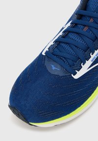 Modrá sportovní obuv s prodyšným svrškem z síťoviny, kontrastními bílými prvky a jasně žlutou podrážkou. Vyznačuje se texturovaným povrchem a strukturovaným systémem šněrování.