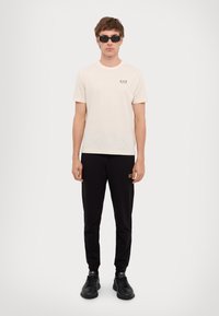 Fehér pamut póló, kis EA7 logóval, fekete melegítőnadrággal, logókiemeléssel, fekete sportcipővel és napszemüveggel párosítva.