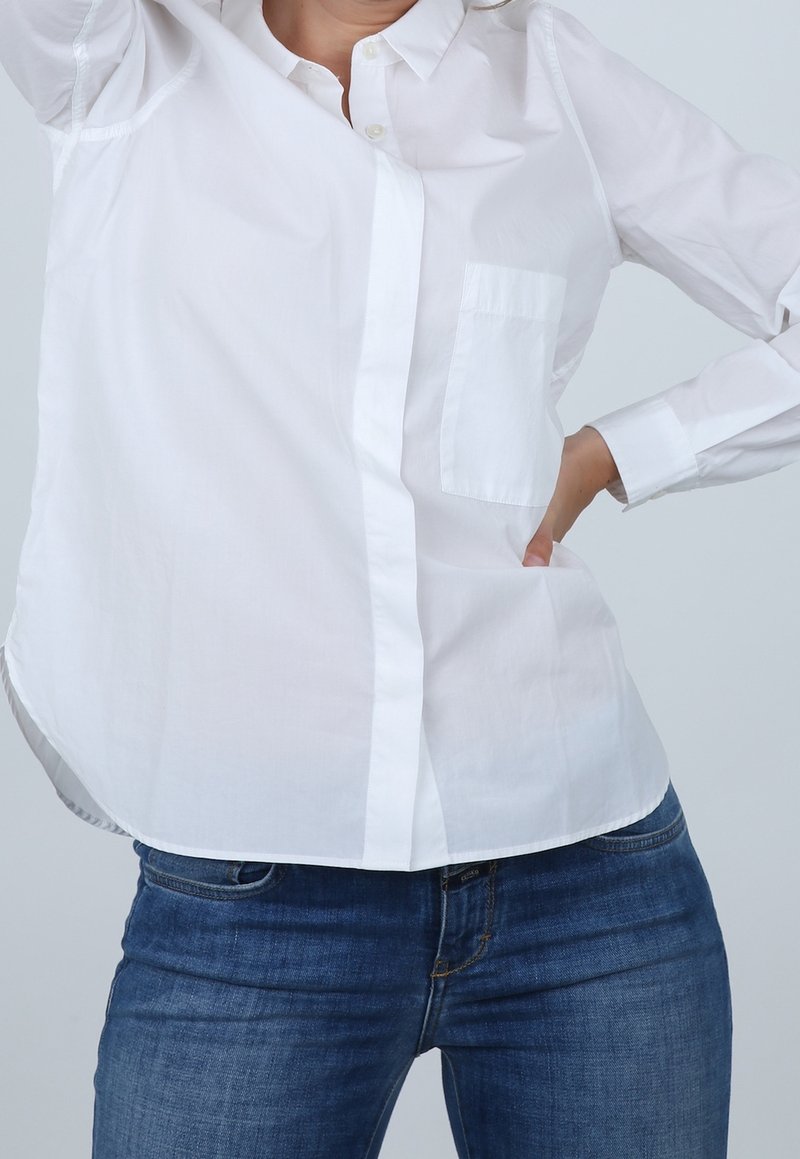 Personne portant une chemise blanche à manches longues boutonnée et un jean bleu, une main sur la hanche et l'autre levée.