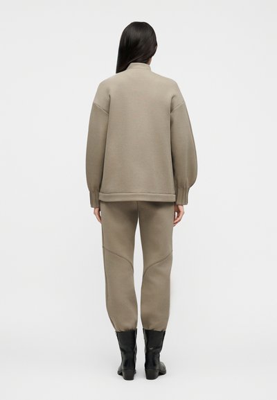 Ženska z dolgimi temnimi lasmi, oblečena v bež prevelik pulover in hlače za tek, stoji obrnjen z zadnje strani, vidni so črni gležnjarji.