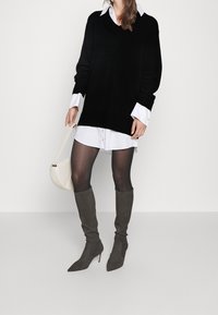 Maglione nero oversize di maglia sopra una camicia bianca con bottoni, abbinato a stivali alti fino alla coscia in suede grigio e a una piccola borsa a spalla bianca.