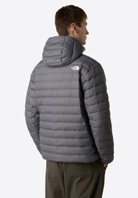 Grå puffjacka med huva, med ett quiltat mönster och elastiska ärmslut. The North Face-logotypen syns på övre ryggen.