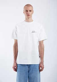 Camiseta de algodón blanca con mangas cortas y escote redondo, que presenta un pequeño logo negro de firma en el pecho, combinada con jeans de color azul claro.
