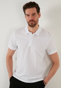 Fehér, texturált anyagból készült pólóing, klasszikus gallérral, rövid ujjakkal és gombos elülső résszel; fekete nadrággal párosítva.