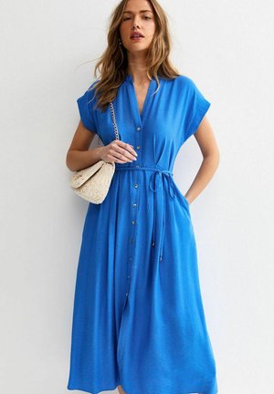 Vrouw die een blauw midi-jurk met knopen draagt, met korte mouwen en een taille met strik, terwijl ze een gebreide crème handtas met kettingriem vasthoudt.