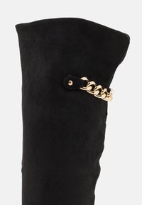 Bota de ante negra hasta la rodilla con la parte superior doblada, que presenta un acento de cadena dorada unida a una correa negra para añadir detalle e interés visual.