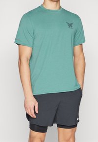 Vaaleansininen lyhythihainen urheilupaita, jossa on teksturoitu kangas. Paidassa on pieni musta logo rinnassa ja kontrastiväriset harmaat yksityiskohdat.