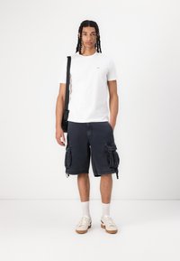Wit T-shirt met ronde hals, donkere cargoshorts met grote zijzakken, witte en beige sneakers, grijze sokken, zwarte schoudertas. Eenvoudig ontwerp.