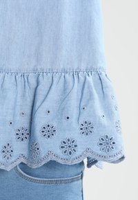 Haut en denim bleu clair avec un ourlet festonné, orné de broderies florales et de petits détails en œillets, ajoutant de la texture et un intérêt au design.