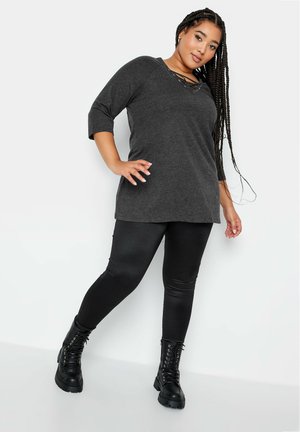Graues Tunika-Top mit ¾-Raglanärmeln und einem Schnürkragen. Kombiniert mit schwarzen Leggings und schwarzen Kampfstiefeln. Glatte Textur.
