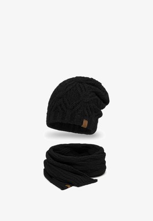 Bonnet en tricot noir avec un motif de tresses complexe et une petite étiquette en cuir, associé à une écharpe en tricot à mailles torsadées assortie. Les deux sont texturés et doux.