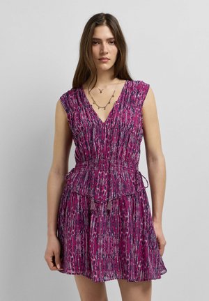 Jeune femme en robe violette à motifs sans manches avec un décolleté en V et une ceinture à la taille, debout devant un fond gris uni.
