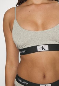Šedý bralet s tenkými ramínkami, ktorý má čierny pás s nápisom "Calvin Klein" a logom "CK". Hladký povrch a jednoduchý dizajn.