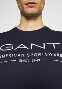 Tummansininen puuvillapaita, jossa on suuri valkoinen printtilogo "GANT AMERICAN SPORTSWEAR" ja "SINCE 1949" etupuolella. Pyöreä kaula-aukko.
