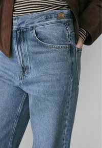 Nahaufnahme einer Person, die hellblaue Jeans, eine braune Jacke und ein gestreiftes Oberteil trägt, während die Hand lässig in der Tasche steckt.