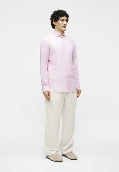 Άνδρας στέκεται φορώντας ένα ανοιχτό ροζ πουκάμισο με κουμπιά, φαρδιά υπόλευκα παντελόνια και μπεζ παπούτσια χωρίς κορδόνια, μπροστά σε απλό φόντο.