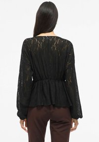 Mustast pitsist pluus pikkade, puhvis varrukatega ja kogutud vöökohaga. Õhuke tekstuur ja lillemuster loovad visuaalse detaili. Sobitatud pruunide pükstega.