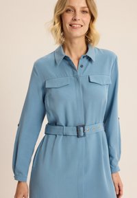Jasnoniebieska sukienka wykonana z gładkiego materiału, z kołnierzykiem, dwoma kieszeniami na piersiach, długimi rękawami oraz ściągniętą talią z dopasowanym paskiem i akcentami ze stali.