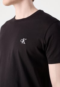 Person wearing a plain black t-shirt with a small white "cK" logo embroidered on the left chest area.