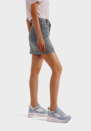 Donna che indossa sneakers azzurro chiaro e bianche, minigonna di jeans e t-shirt bianca, in piedi contro uno sfondo semplice.