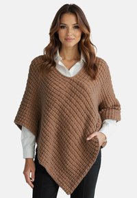 Poncho intrecciato marrone con un motivo testurizzato diagonale, scollo a V, maniche arrotolate e una tasca frontale, indossato sopra una camicia bianca.