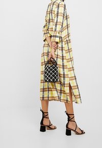 Żółta sukienka w kratę o długości do kolan i bocznym rozcięciu, zestawiona z czarnymi sandałami na obcasie i małą czarno-białą torebką.