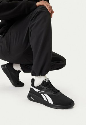 Persona con zapatillas Reebok en blanco y negro, calcetines blancos y pantalones negros, agachada con la mano apoyada en la rodilla.
