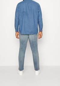 Man standing facing away wearing light blue jeans, a blue denim shirt, white sneakers, and white socks against a plain white background.