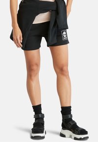 Shorts en coton noir avec un écusson carré du logo Timberland, assortis à des sandales noires épaisses et des chaussettes noires texturées.