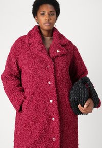 Abrigo borroso de color burdeos con cuello con muescas y botones a presión. El modelo sostiene un bolso negro texturizado, que presenta un asa en forma de lazo.