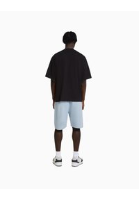 Muž s pletenými vlasy, oblečený v volném černém tričku, světlomodrých kraťasech po kolena, bílých ponožkách a černobílých teniskách, stojící zády.