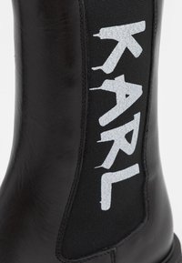 Black leather boot featuring a stretch fabric panel with prominent white "KARL" text, smooth texture, rounded toe, and flat sole.