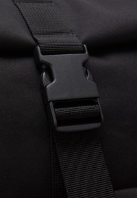 Musta muovinen sivulukollinen solki, kiinnitetty mustaan kudottuun hihnaan, teksturoidun mustan kangaspinnan päällä.