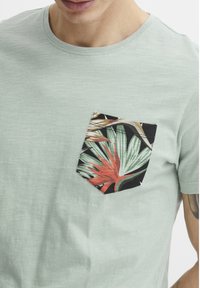 Camiseta de manga corta verde claro hecha de una tela suave, con un bolsillo tipo parche negro que presenta un vibrante patrón de hojas tropicales en verde y rojo.