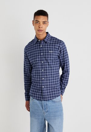 Camisa de cuadros marrón marino con patrón de cuadrícula blanca, de manga larga y tejido suave. Presenta un logo en el pecho izquierdo y un cuello clásico.