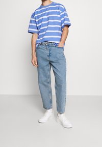 Blå och vit randig t-shirt i bomull, matchad med ljusblå denimjeans. Modellen har vita sneakers och händerna i fickorna.