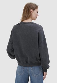 Frau mit hellbraunem Haar, die ein dunkelgraues, übergroßes Sweatshirt und blaue Jeans trägt, von hinten vor weißem Hintergrund gezeigt.