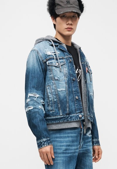 Νέος άνδρας που φοράει ένα φθαρμένο τζιν μπουφάν πάνω από μια γκρι φούτερ με κουκούλα, συνδυασμένο με μπλε τζιν και σκουρόχρωμο καπέλο του μπέιζμπολ.