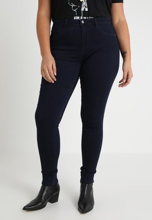 Woman wearing dark blue skinny jeans and black heeled ankle boots, standing against a plain light background.