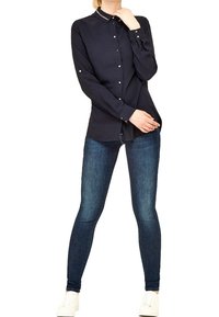 Mujer vestida con una camisa negra de botones, jeans ajustados oscuros y zapatillas blancas, de pie con una mano cerca de su cuello y la otra en la cadera.