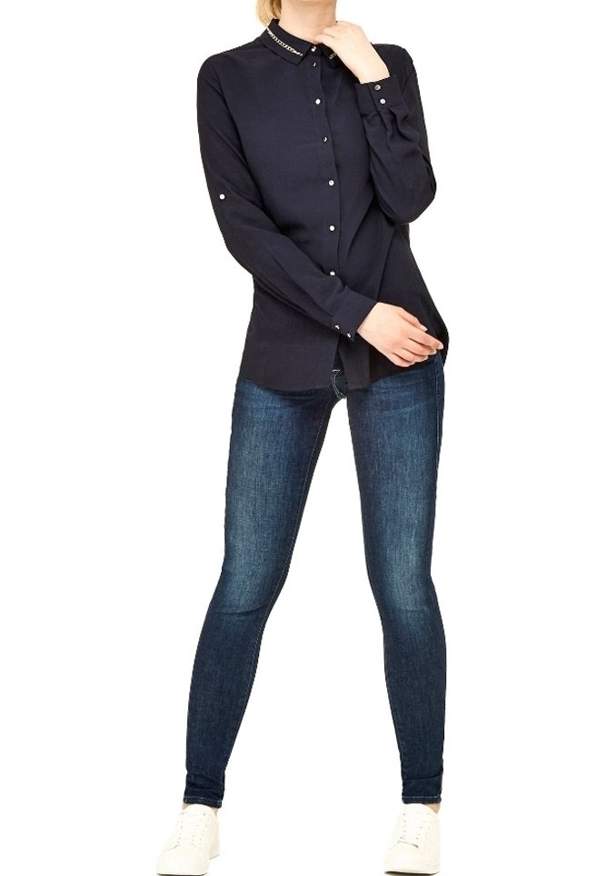 Mujer vestida con una camisa negra de botones, jeans ajustados oscuros y zapatillas blancas, de pie con una mano cerca de su cuello y la otra en la cadera.