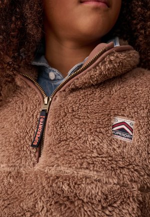 Kiharatukkainen henkilö, joka pukeutunut vaaleanruskeaan pörröiseen Superdry Mountain Sports -vetoketjutakkiin sinisen napitetun paidan päällä.
