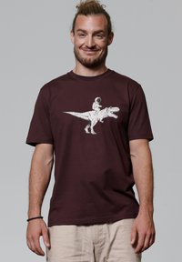 Bordeaux katoenen t-shirt met een witte afbeelding van een dinosauruss met een ruimtepak figuur dat er op rijdt, ronde hals en ontspannen pasvorm.