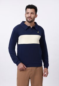 Man wearing navy blue long-sleeve polo with white horizontal stripe and tan pants, standing against a white background, looking right.