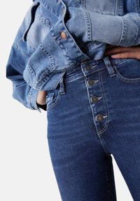 Tummansiniset korkeavyötäröiset farkut viidellä nappisuljennalla, joissa on sileä pinta ja kevyt joustavuus, yhdistettynä vaaleansiniseen farkkutakkiin.