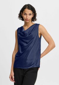 Top sin mangas de satén azul marino con escote drapeado, textura suave y corte relajado. Combinado con pantalones negros, que destacan un diseño minimalista.