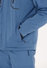 Person trägt blaue Jacke und Hose, eine Hand in der Jackentasche, zeigt Details der Reißverschlusstasche an der Oberbekleidung.