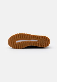 Black sneaker sole with a textured, ridged design. Made of rubber with a brown, wavy tread pattern and a circular logo at the heel.