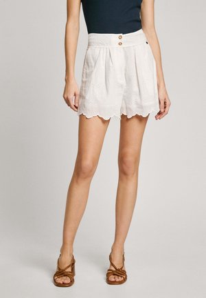 Frau trägt weiße, hoch taillierte Lochspitzen-Shorts mit gewelltem Saum und braune, riemchige Absatzsandalen, stehend vor einfarbigem Hintergrund.