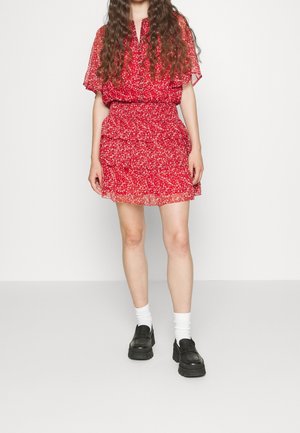 Frau trägt ein kurzes rotes Blumenkleid mit flatternden Ärmeln, weißen Socken und schwarzen klobigen Loafers, steht vor einem schlichten Hintergrund.